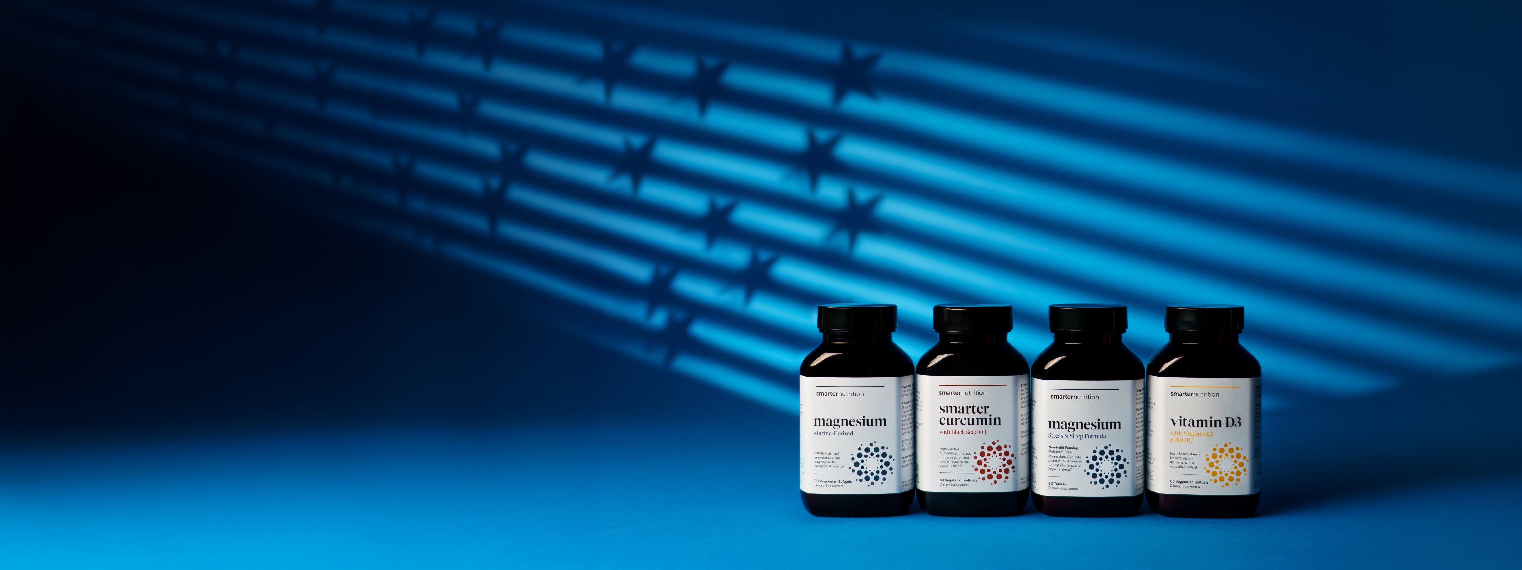 Four dark brown supplement bottles from Smarter Nutrition are lined up on a vibrant blue surface. The bottles feature clean white labels with modern minimalist typography and colorful circular, dot-matrix geometric patterns. From left to right, the labels read "magnesium," "smarter curcumin," "magnesium," and "vitamin D3." A dramatic, stylized blue light casts horizontal stripes and subtle star shapes across the background behind the products.