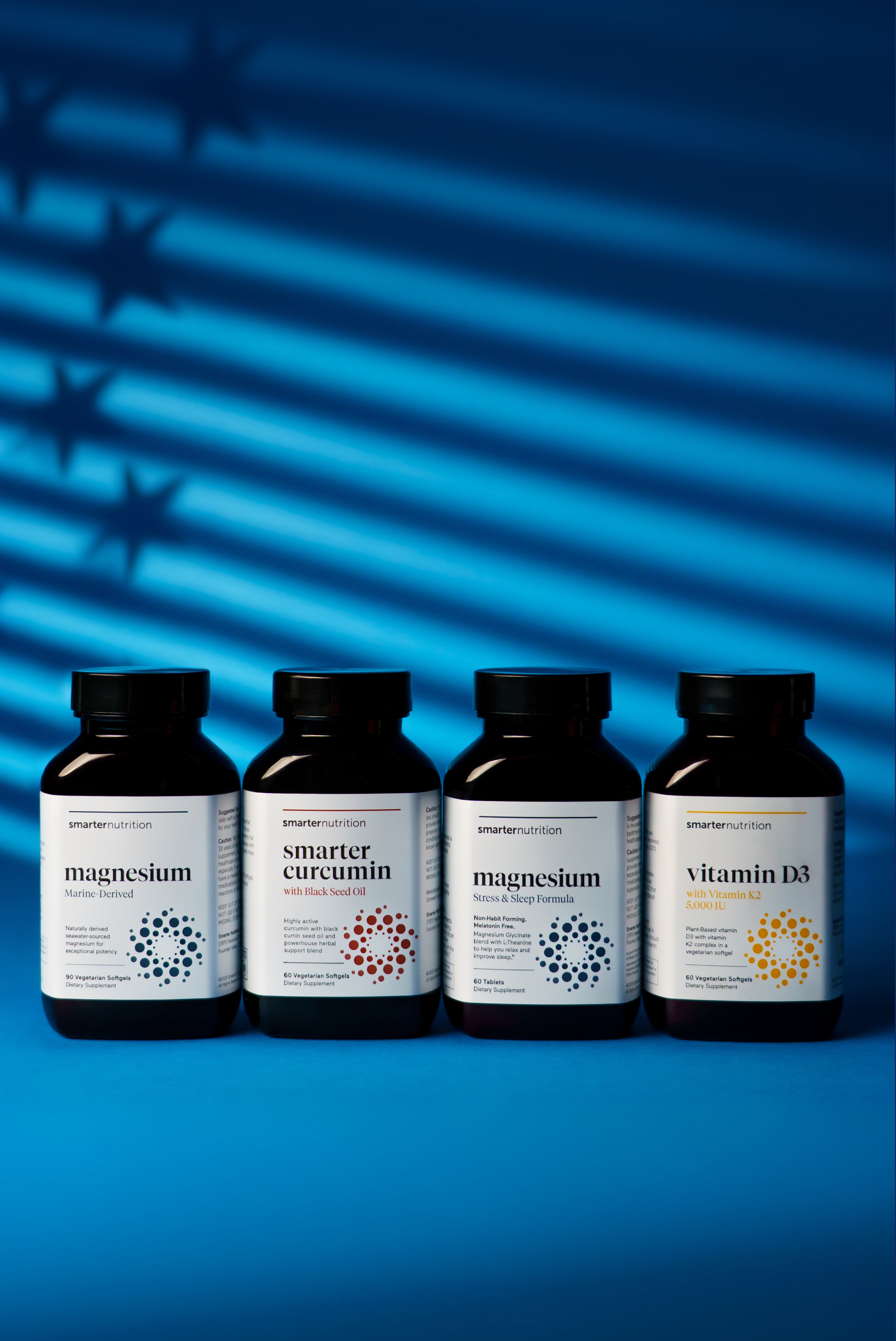 Four dark brown supplement bottles from Smarter Nutrition are lined up on a vibrant blue surface. The bottles feature clean white labels with modern minimalist typography and colorful circular, dot-matrix geometric patterns. From left to right, the labels read "magnesium," "smarter curcumin," "magnesium," and "vitamin D3." A dramatic, stylized blue light casts horizontal stripes and subtle star shapes across the background behind the products.