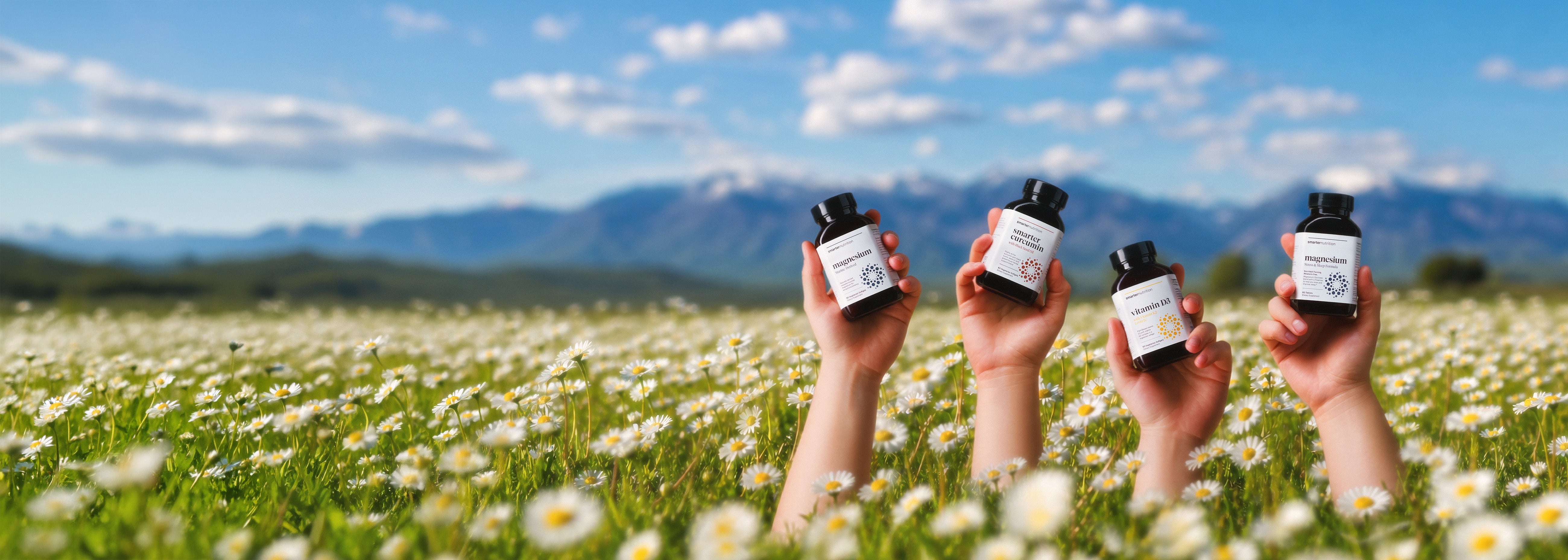 Four hands holding small white bottles with black caps above a field of white flowers and mountains under a blue sky.